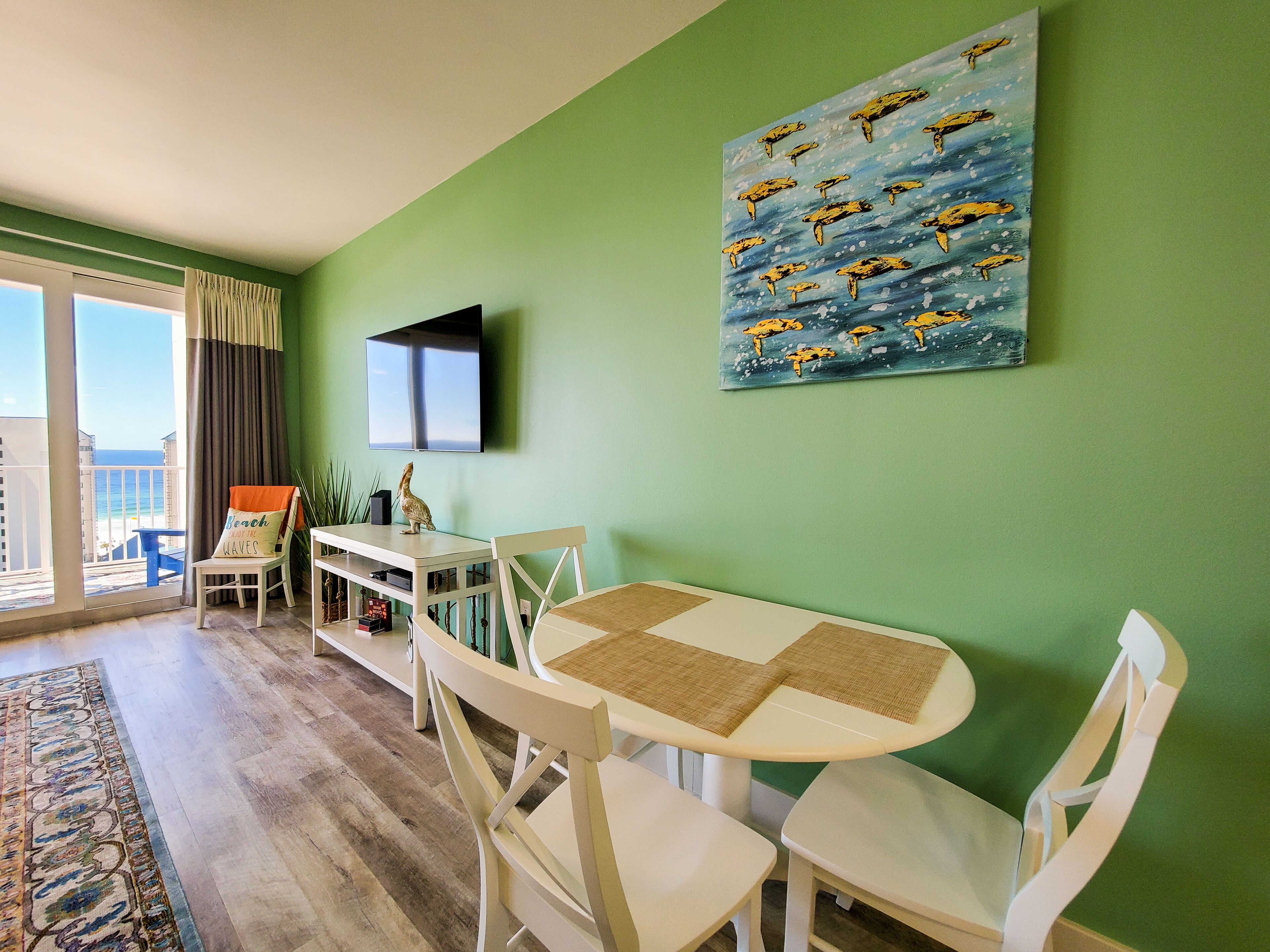 A room with a green wall, a flat-screen TV, a painting of dolphins, a dining table with chairs, a small table with a speaker, and a view of the ocean through a balcony.