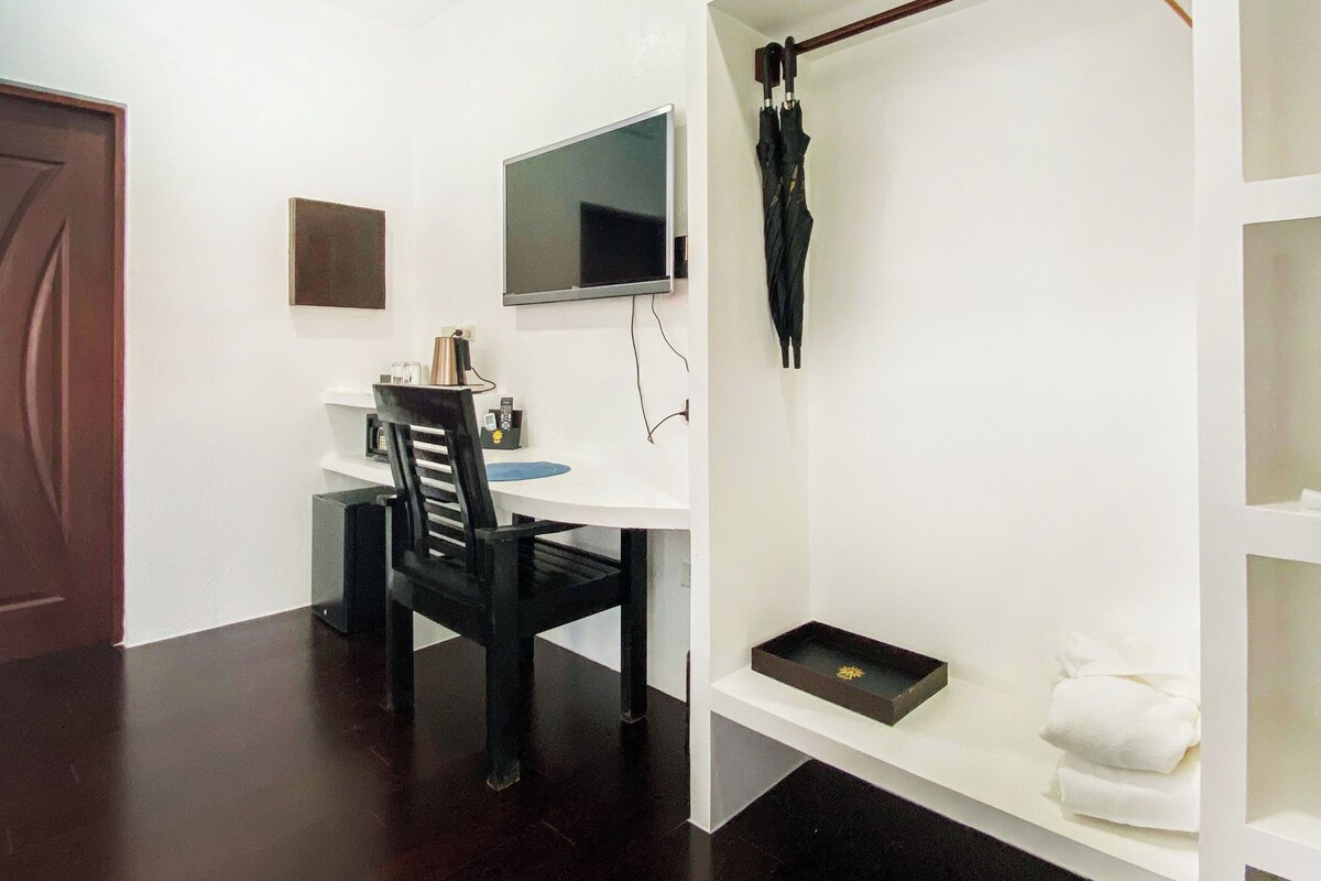 A room with a dark wooden floor, a white desk with a chair, a mounted TV, and a shelf with a black tray and white towels.