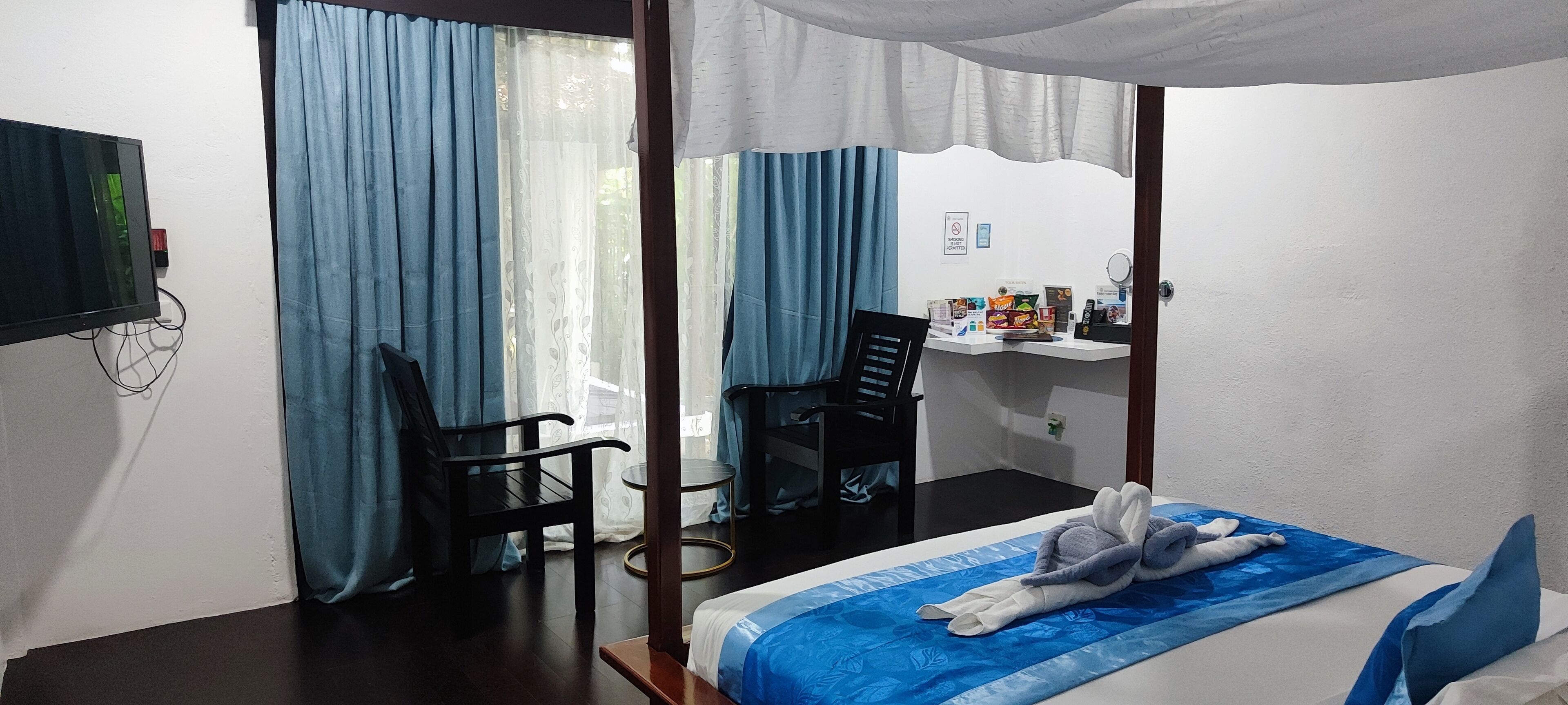 Una habitación con cama, escritorio, dos sillas, televisión y ventana con cortinas.