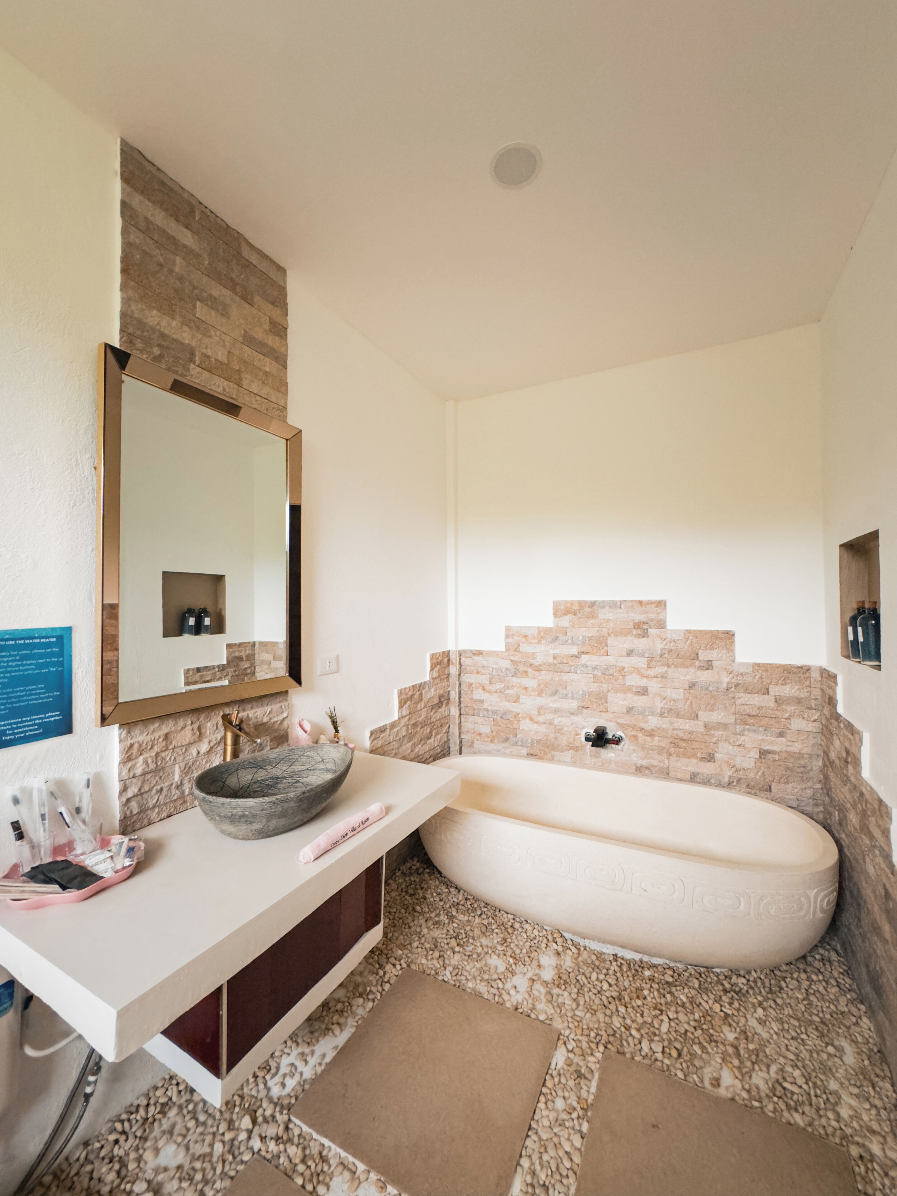 Une salle de bain avec un lavabo blanc, un grand miroir, un mur en pierre et une baignoire.