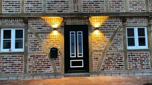 A traditional brick building with a black door, two lit windows, and a wooden staircase. - New charming half-timbered house by the lake in the idyllic Münsterland region (Warendorf)