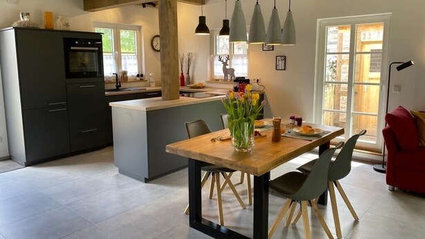 A modern kitchen with a central island, pendant lights, and a dining area with a wooden table and chairs. - New charming half-timbered house by the lake in the idyllic Münsterland region (Warendorf)