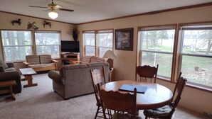 A cozy living room with a sofa, chairs, a TV, and large windows offering a view of the outdoors. - Cabin Getaway! (Remer)