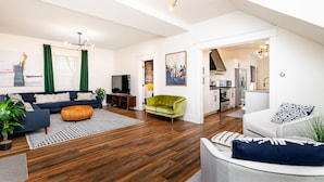 A modern living room with a sofa, a green armchair, and a dining area with a kitchen in the background.