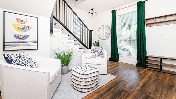 A modern living room with a staircase, white furniture, a green plant, and a framed artwork on the wall.