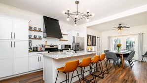 A modern kitchen with white cabinets, a central island with bar stools, a large flat-screen TV, and a dining area with a table and chairs.