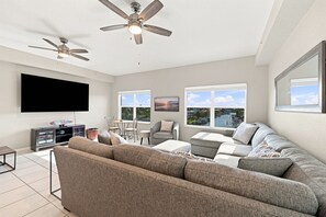 A modern living room with a sectional sofa, a flat-screen TV, ceiling fans, and large windows offering a view of a marina.