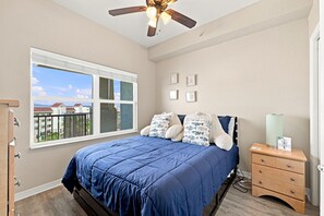 Quarto com cama, mesa de cabeceira, cômoda, ventoinha no teto e janela com vista para edifícios.