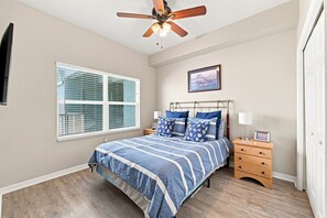 A bedroom with a bed, a ceiling fan, a window with blinds, a bedside table with a lamp, and a framed picture on the wall.