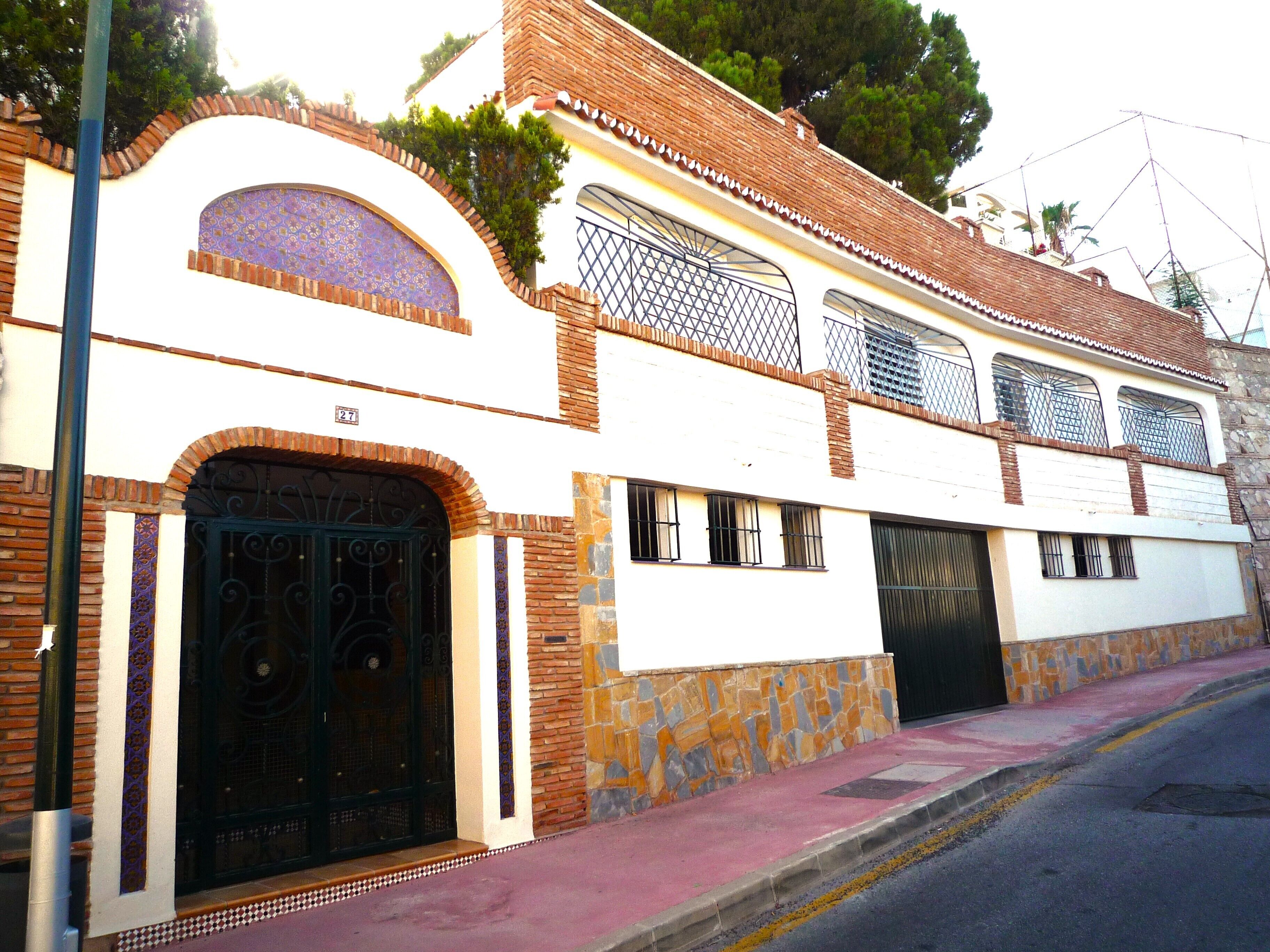 Un edificio de dos pisos con fachada blanca, una reja negra y un muro de piedra.