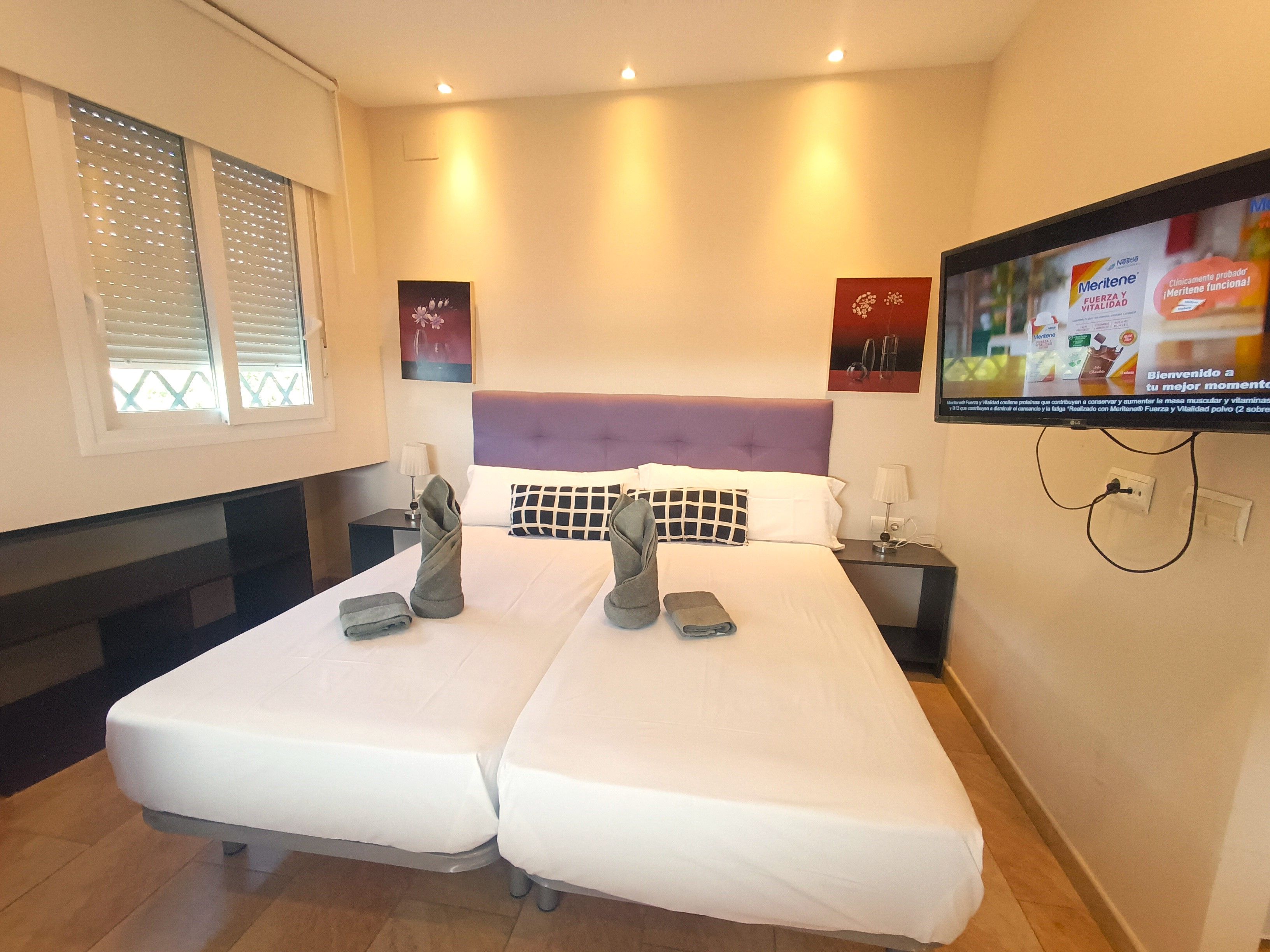 A hotel room with two beds, a television mounted on the wall, and a window with blinds.