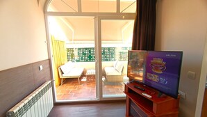 Een kamer met een schuifdeur die uitkomt op een buitenpatio. Er is een televisie op een houten standaard, een radiator aan de muur en een kleine tafel met een stoel.