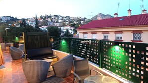 A terrace with wicker furniture, a glass table, and a view of a town with buildings and a mountain. - D-2 Apartment with a terrace. (Malaga)