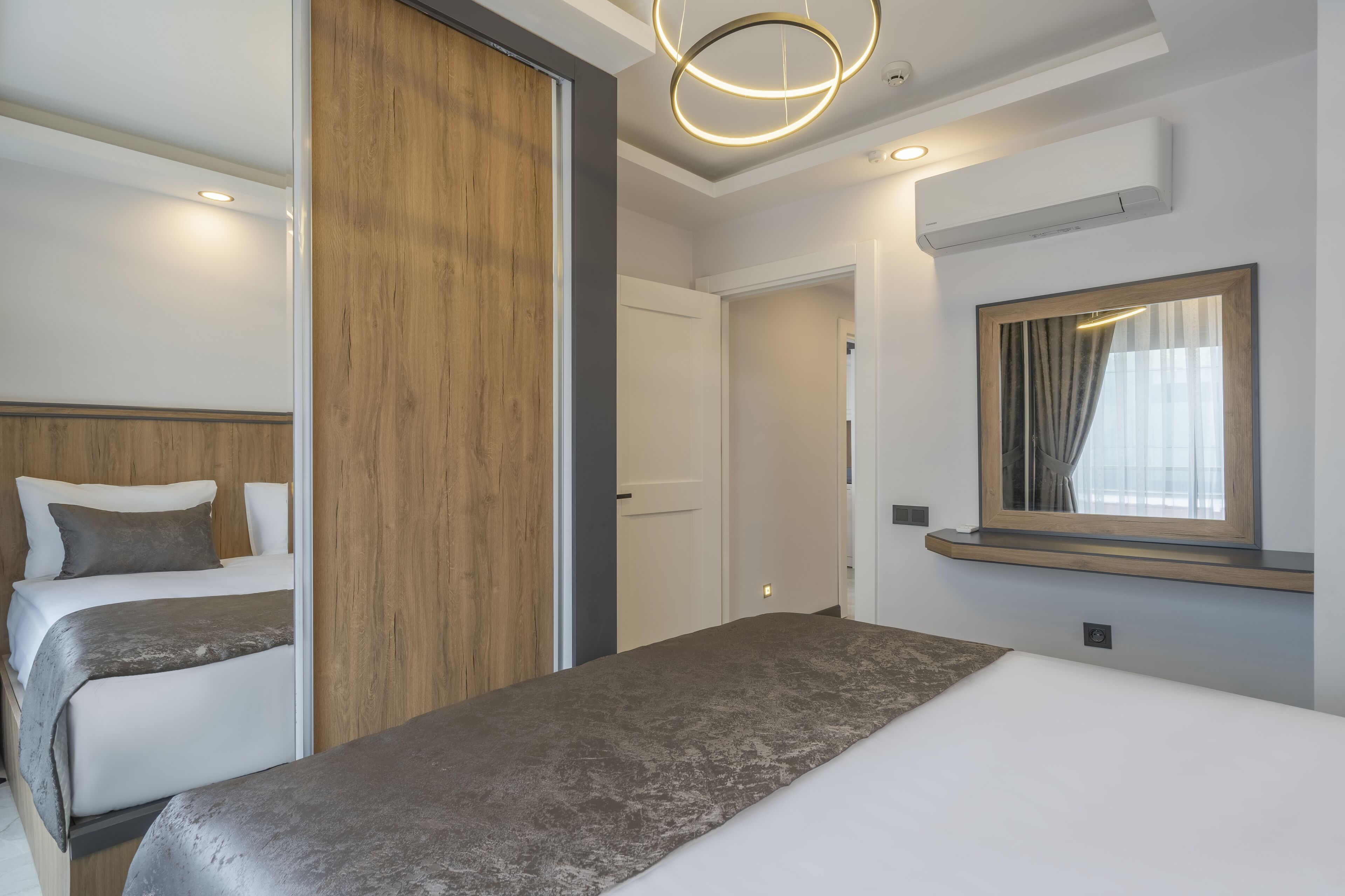 A hotel room with two beds, a wooden panel wall, a large mirror, and a modern light fixture.