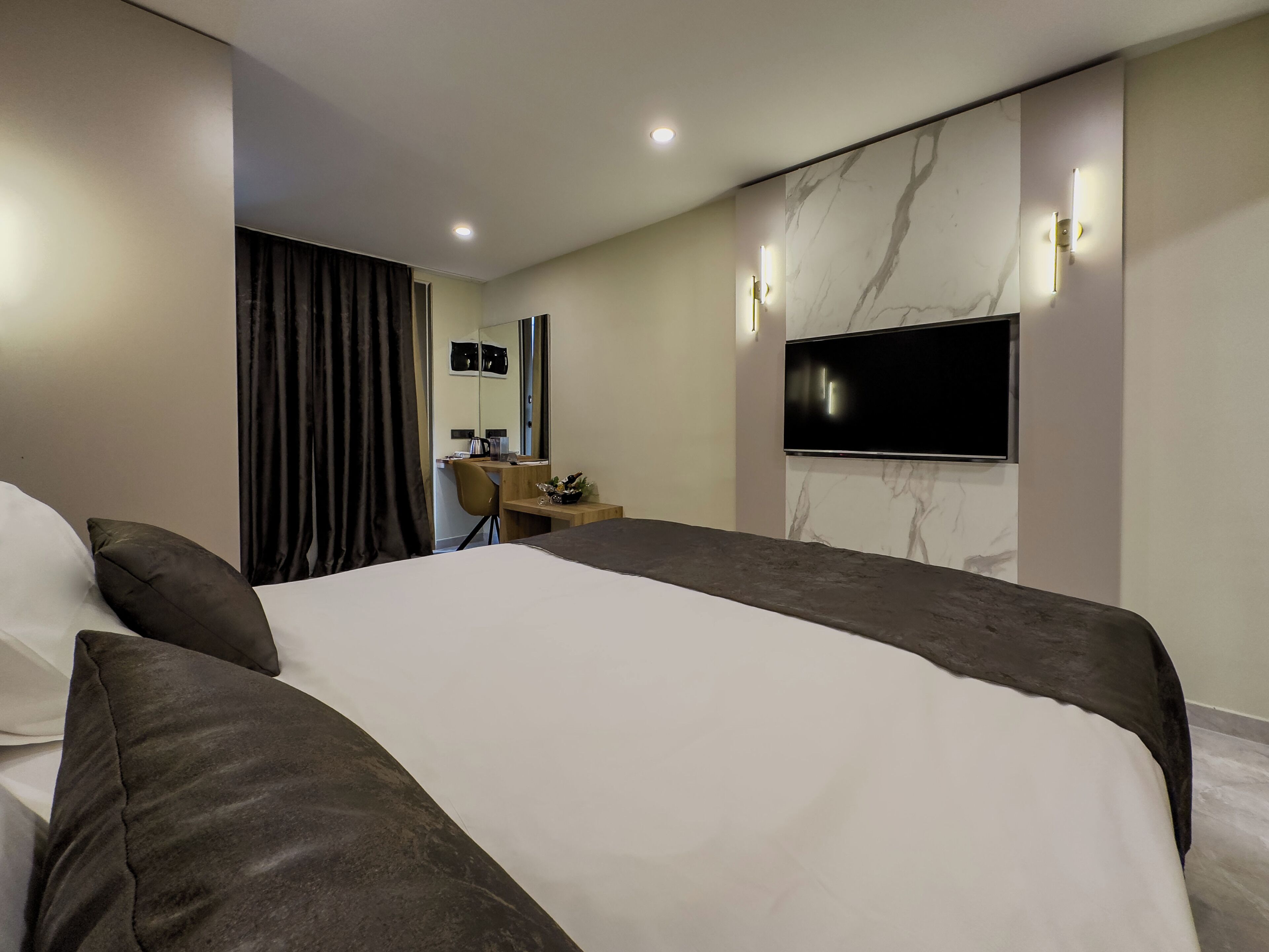 Una habitación de hotel moderna con una cama grande, un escritorio, una silla, un televisor de pantalla plana montado en la pared y un espejo.
