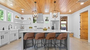 A modern kitchen with white cabinetry, a central island with bar stools, pendant lighting, and a view to an outdoor area. - Broken Bow Secluded Playset Retreat for Families (Broken Bow)