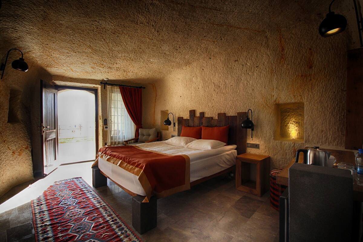 A bedroom with a stone ceiling, a bed with a red and white bedspread, a bedside table with a lamp, and a view of the outside through a door.