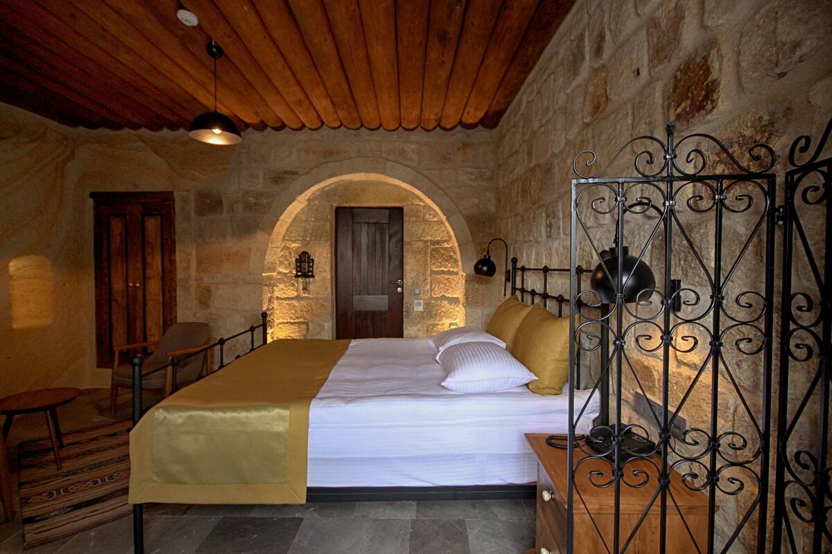A bedroom with a stone wall, wooden ceiling, a bed with white bedding, a wooden chair, and a metal gate.
