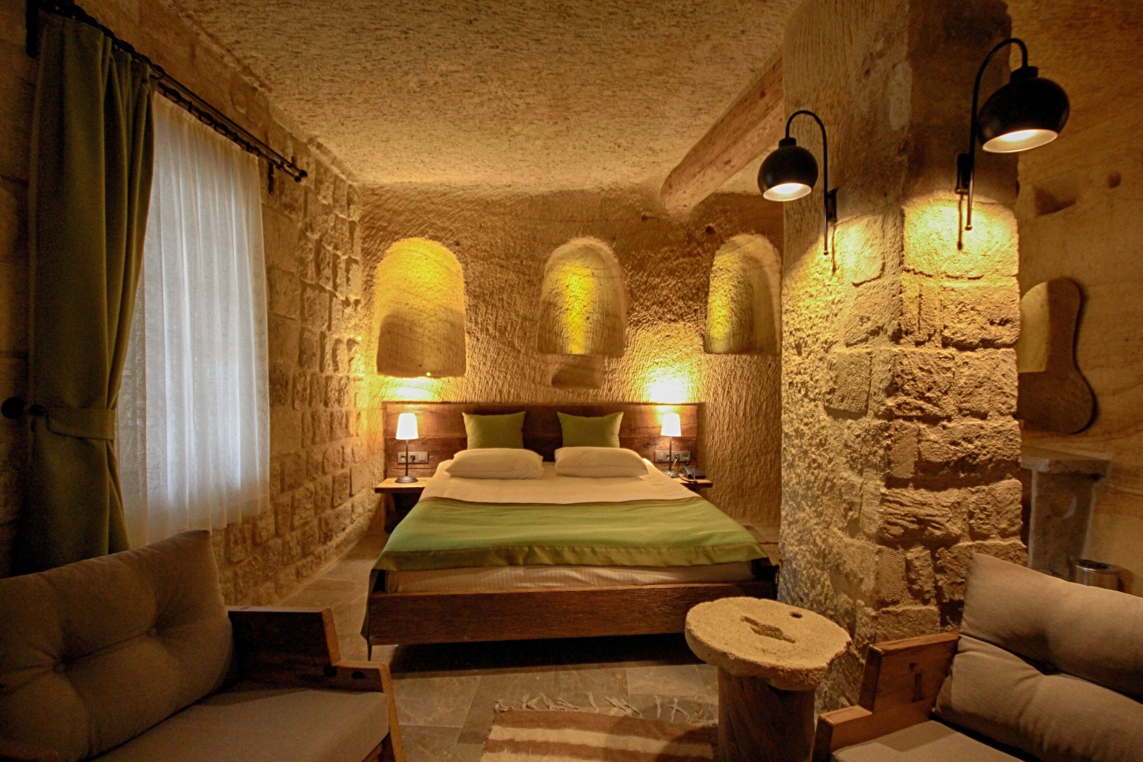 Un dormitorio acogedor con paredes de piedra, una cama, dos mesitas de noche con lámparas, un taburete redondo de madera y un sofá.
