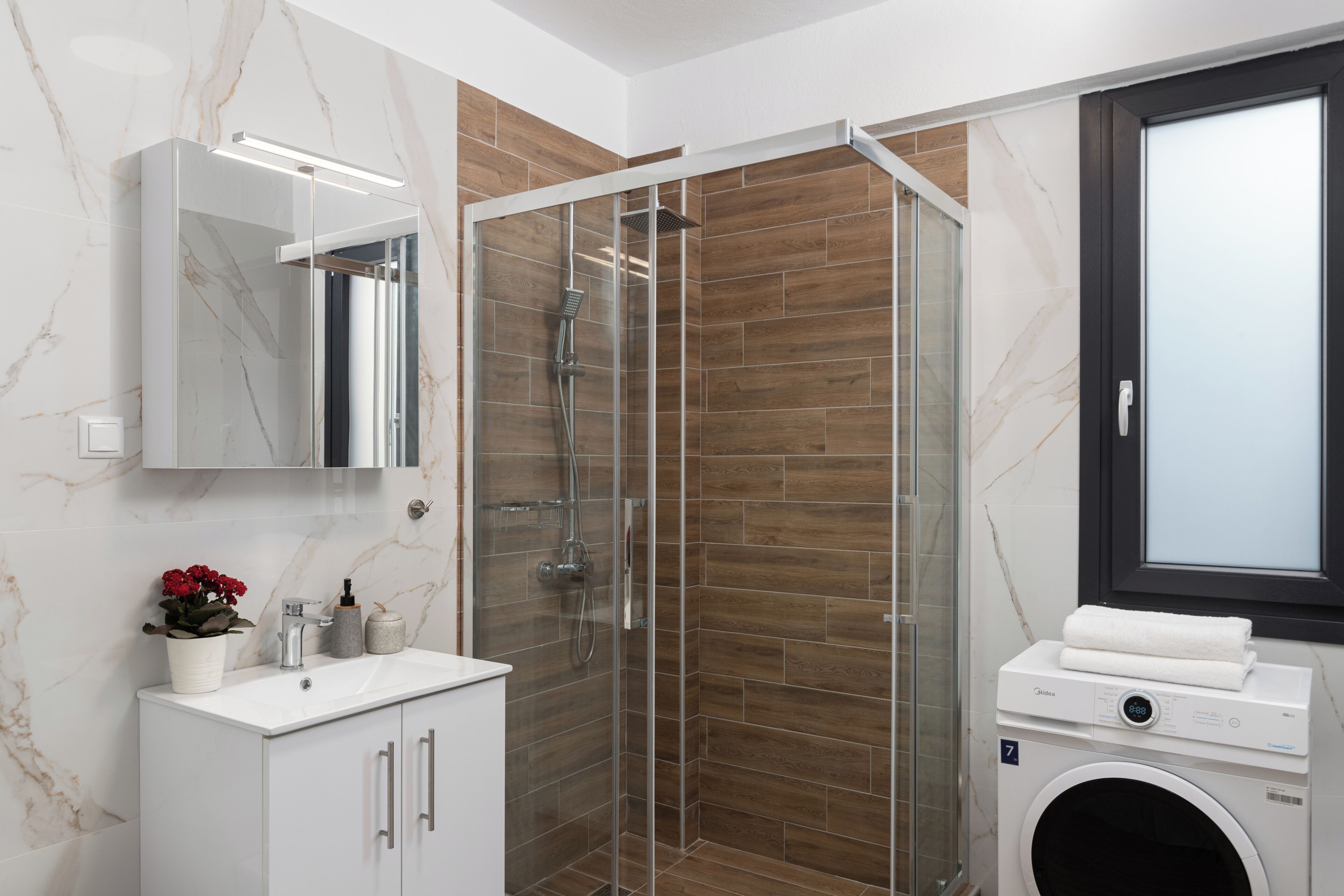 A modern bathroom with a glass-enclosed shower, a white sink with a marble backsplash, and a built-in washing machine.