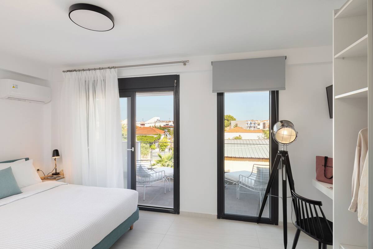 Habitación de hotel con cama, escritorio, silla, lámpara, ventana y balcón con mobiliario exterior.