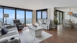 A spacious living room with a large window overlooking the sea, a grey sofa, a white coffee table, and a dining area with wicker chairs.