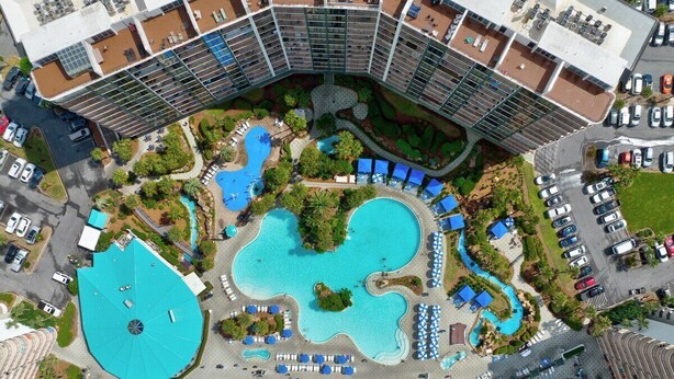 An aerial view of a hotel with multiple swimming pools, a large central pool, and a surrounding landscaped area with lounge chairs and umbrellas.