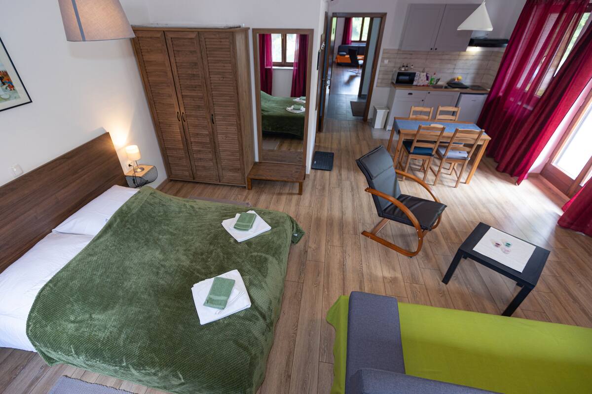 A hotel room with a bed, a green sofa, a dining table with chairs, a kitchen area, and a bathroom visible through an open door.