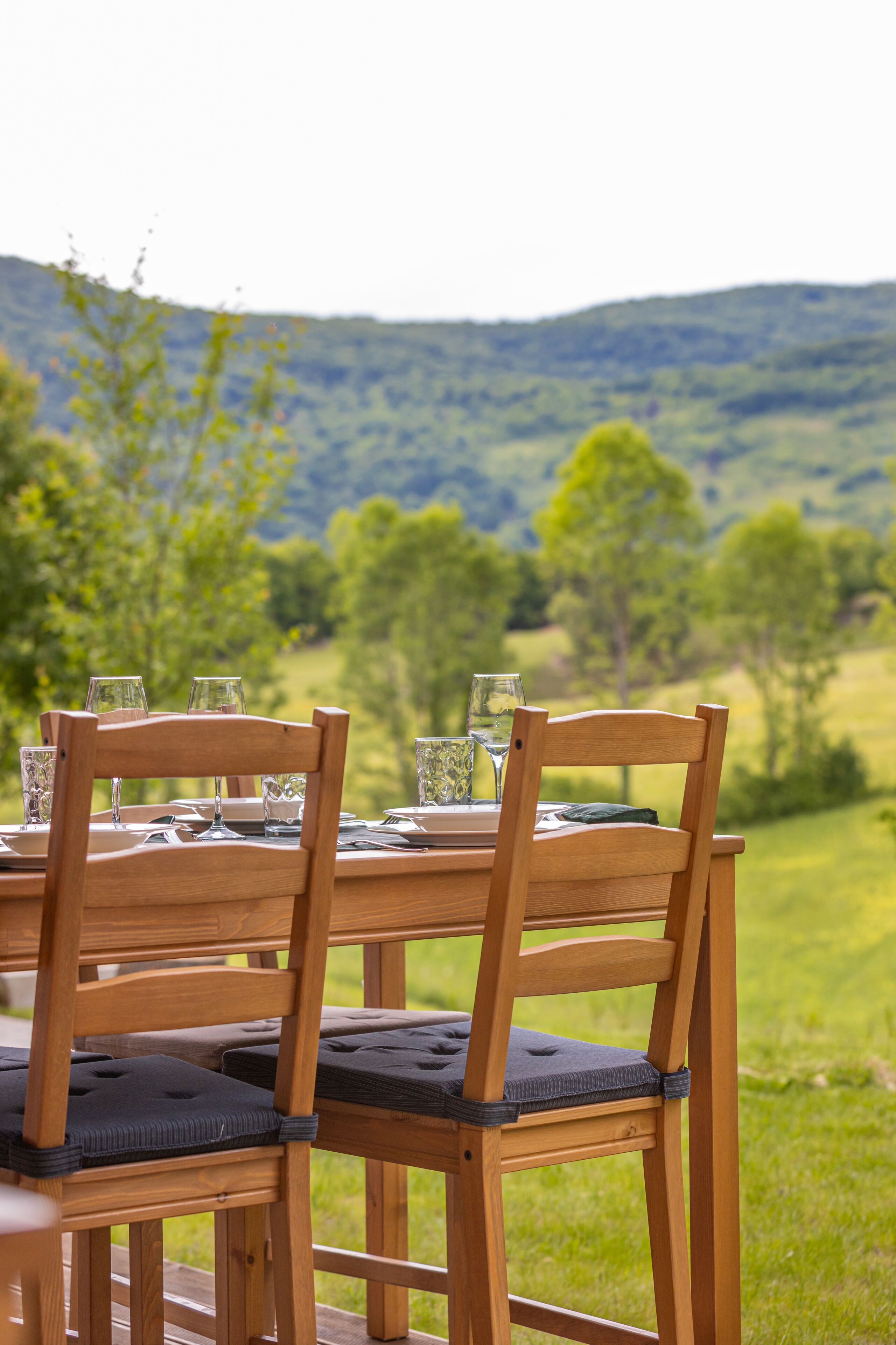 Une table à manger en bois, avec des chaises et des verres, est installée à l’extérieur, offrant une vue sur un paysage verdoyant et vallonné.