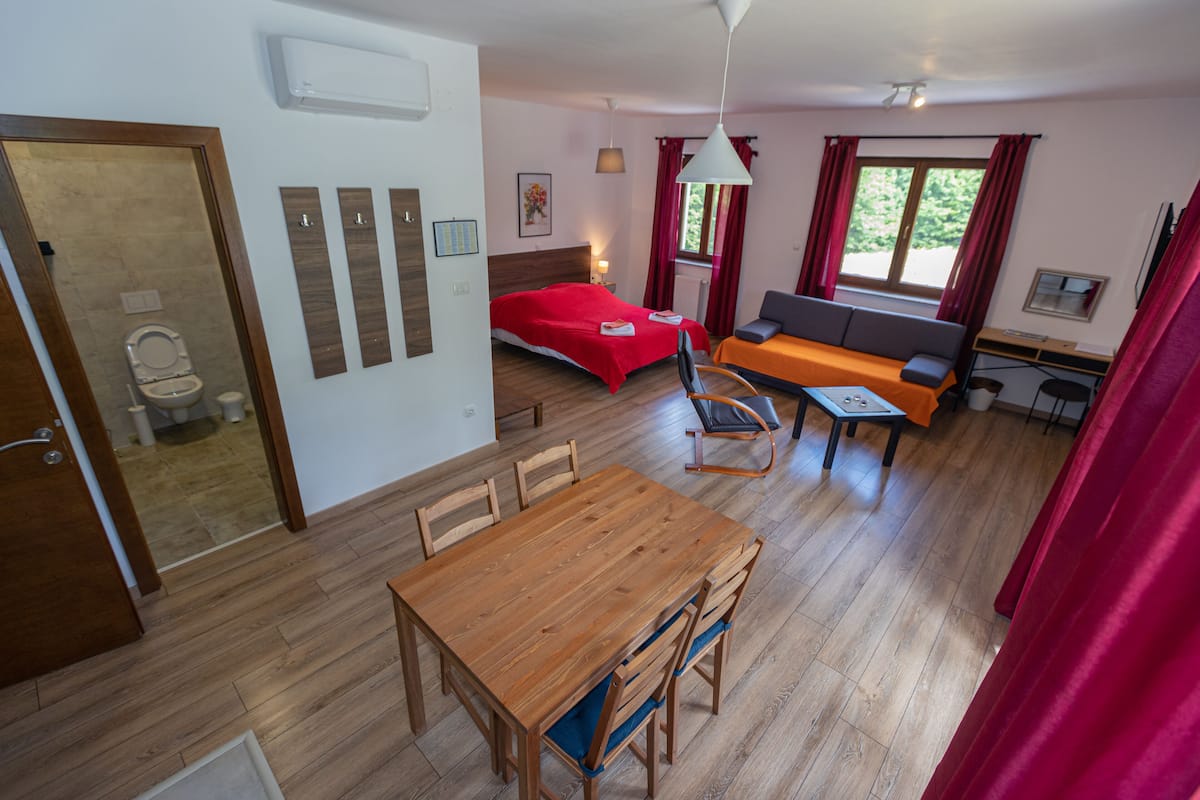 A room with a wooden dining table, a bed with red bedding, a sofa, and a desk.