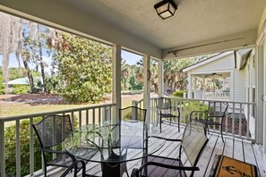 A covered porch with a glass table and chairs, surrounded by a white railing, overlooking a garden and a house.
