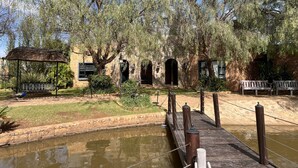 A rustic building with arched windows and a brick facade, surrounded by lush greenery and a wooden bridge over a pond.