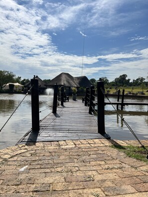 A wooden bridge over water with a thatched-roof structure in the background. - Swan Lake Suites  (Benoni)