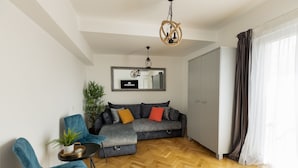 Ein modernes Wohnzimmer mit einem grauen Ecksofa, türkisfarbenen Sesseln, einem kleinen runden Tisch mit einer Pflanze und einem Holzboden mit Fischgrätmuster.