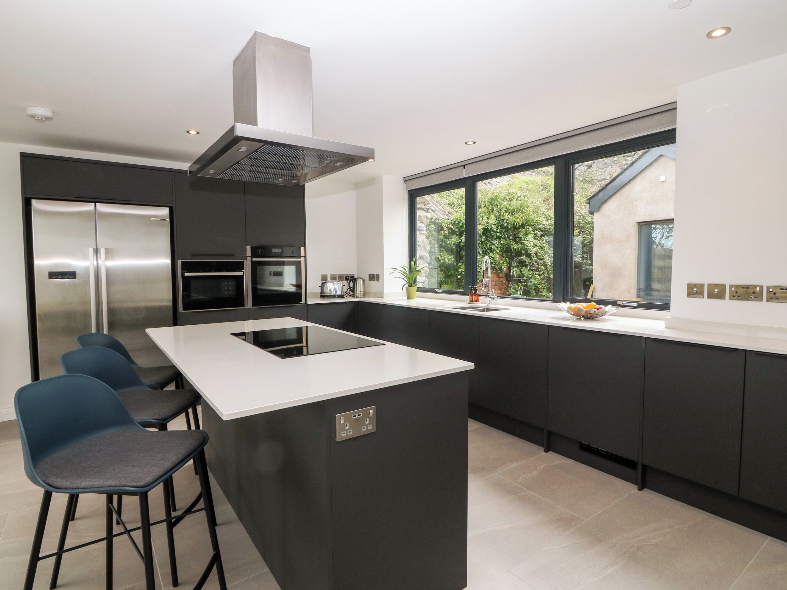 A modern kitchen with a central island, bar stools, a stainless steel refrigerator, built-in oven, and a large window offering a view of greenery.