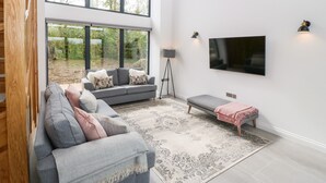 Un salon spacieux avec une grande fenêtre, un canapé gris, un petit banc et un téléviseur à écran plat fixé au mur.