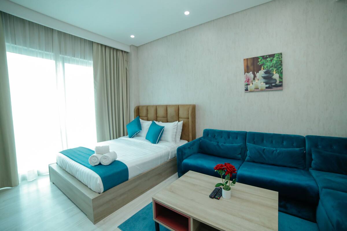 A modern hotel room with a bed, a blue sofa, a wooden coffee table, and a painting on the wall.