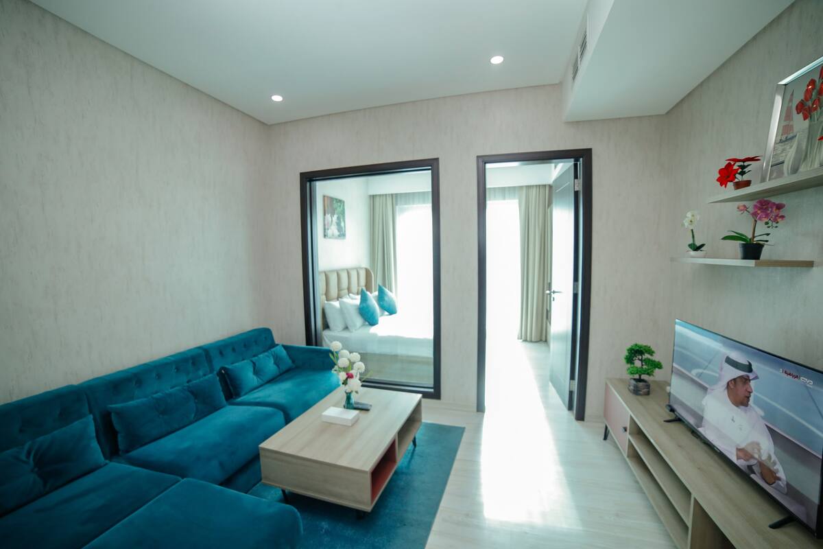 A modern living room with a teal sofa, a wooden coffee table, and a flat-screen TV. There is a large mirror reflecting the room and a small potted plant on the coffee table.