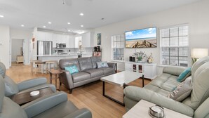 Ein modernes Wohnzimmer mit grauem Sofa, einem Couchtisch und einem Fernseher. Im Hintergrund ist eine Küche mit weißen Schränken und einem Kühlschrank zu sehen.