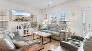 Ein geräumiges Wohnzimmer mit großem Fenster, einer Couch, einem Couchtisch und einem Fernseher, der eine Strandansicht zeigt.