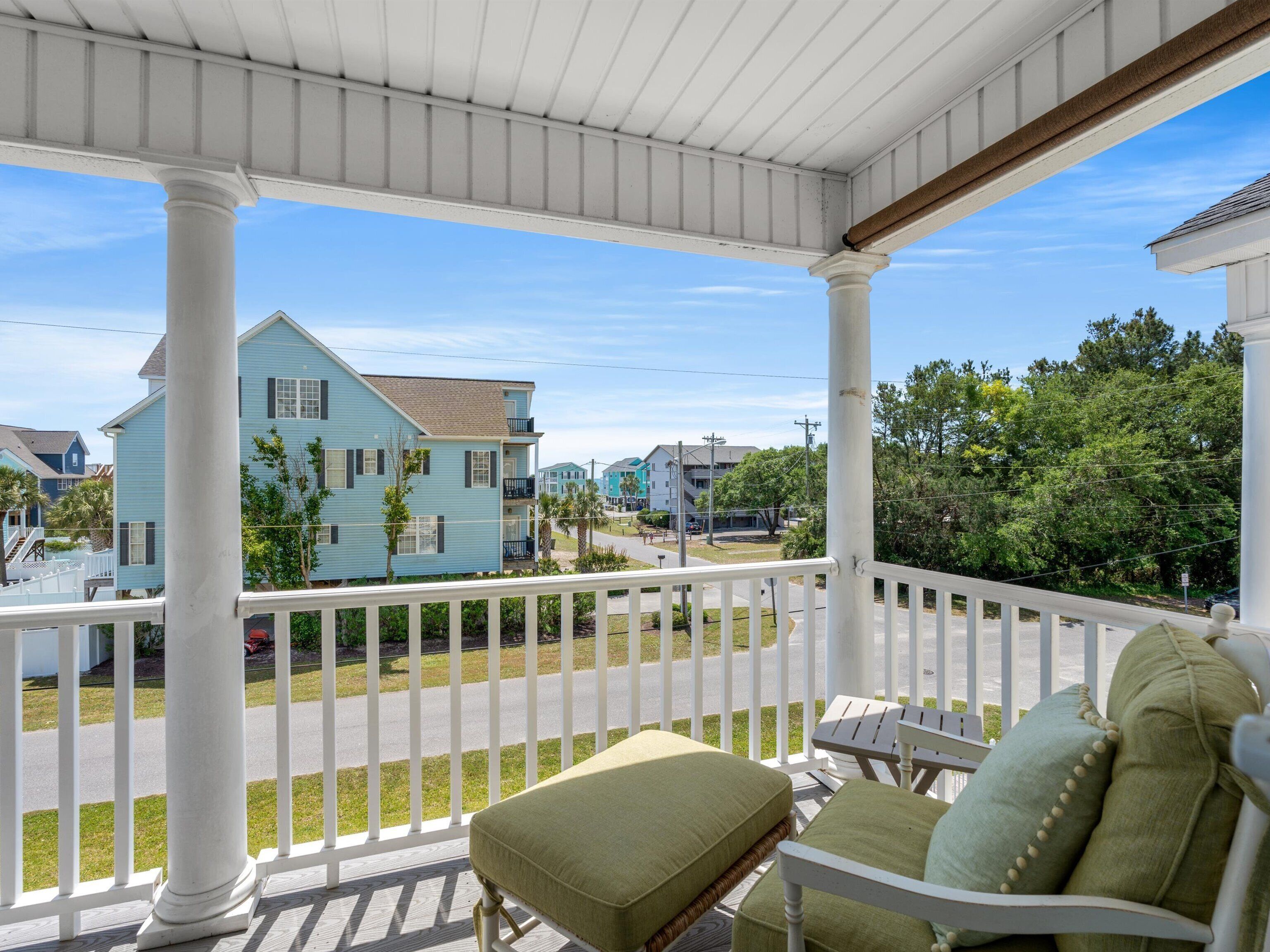 A porch with white railings, a green cushioned bench, and a view of a residential area with houses and trees.