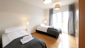 A hotel room with two beds, wooden flooring, a large window with curtains, and a door with a handle. - The Private Apartments (Borgarnes)