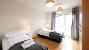 A hotel room with two beds, wooden flooring, a large window with curtains, and a door with a handle.