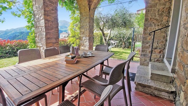 Un espacio exterior de comedor con arcos de piedra, una mesa y sillas de madera, rodeado de follaje y vistas a colinas lejanas.