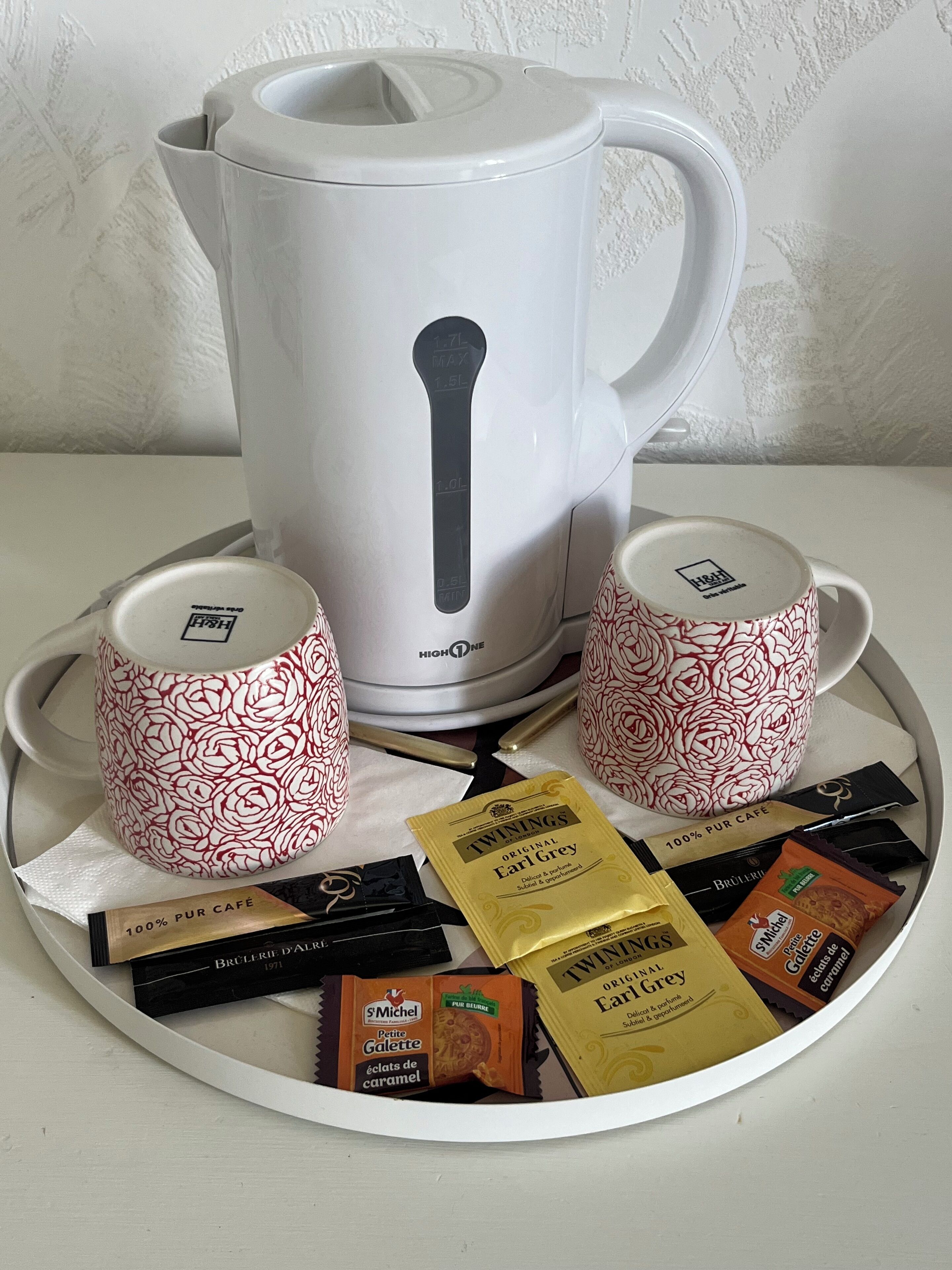 A white coffee maker with a black spout, two matching white cups with red floral patterns, and a tray with various coffee packets.