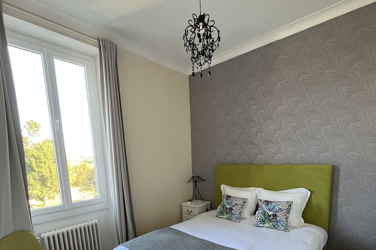 Une chambre à coucher avec un grand lit, une tête de lit verte, une table de chevet, une lampe, une chaise et une fenêtre avec des rideaux.