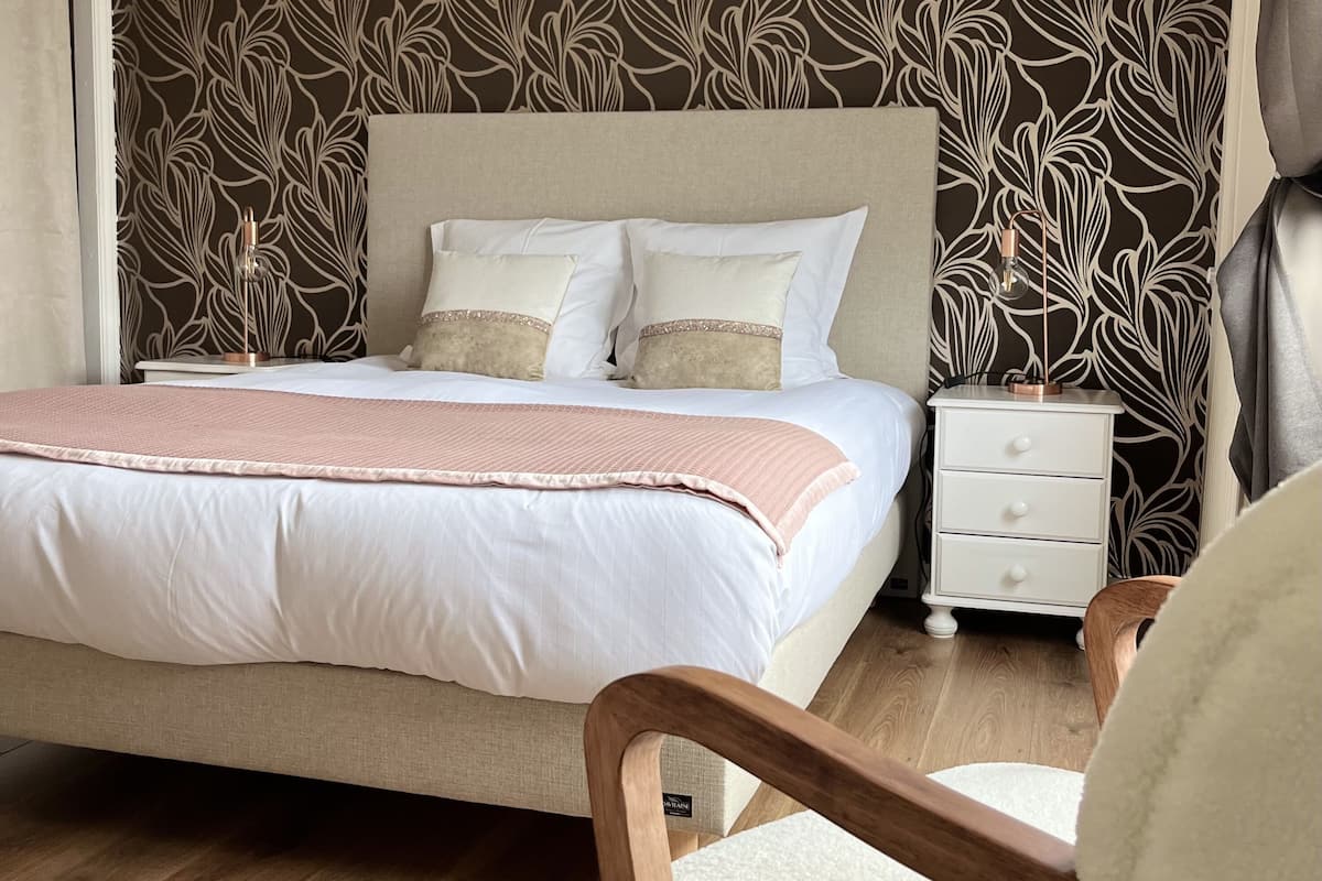 Un lit bien fait, recouvert d’une couverture rose, une table de chevet en bois avec des tiroirs et une chaise dotée d’un coussin blanc.