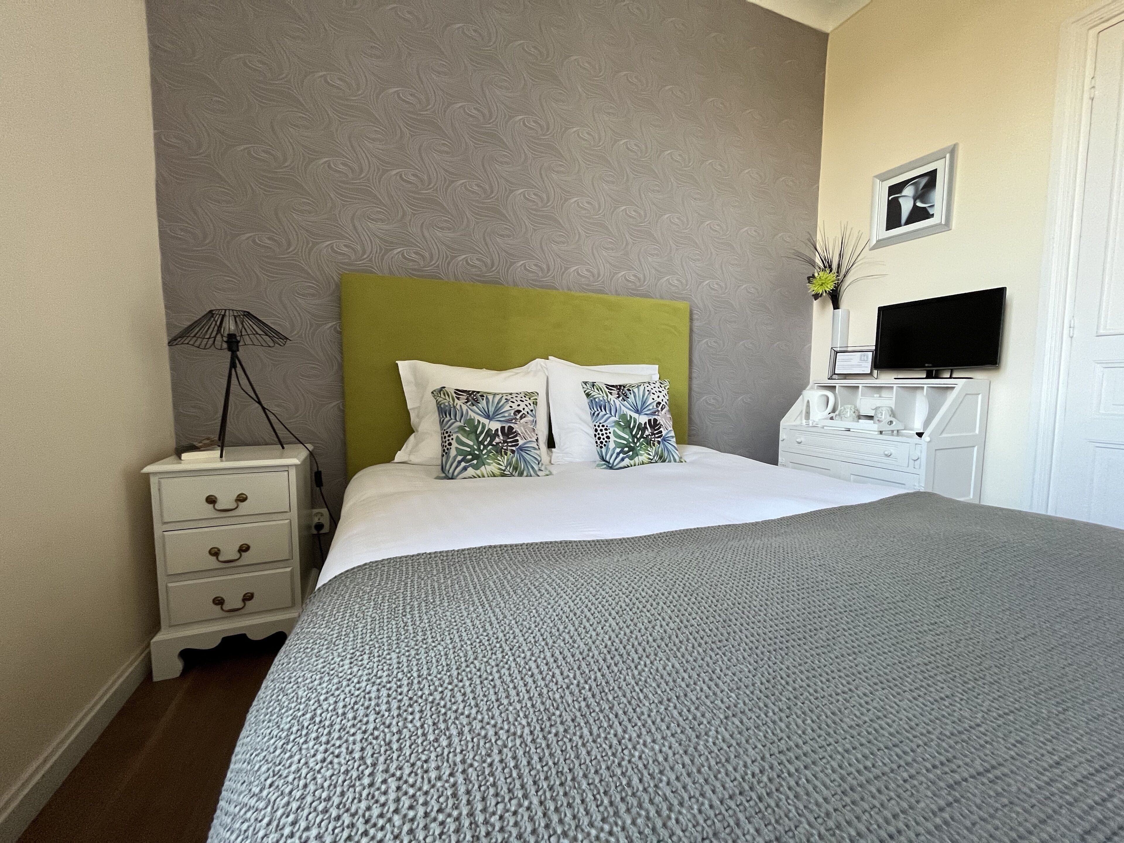 A neatly made bed with a grey knitted blanket, a green headboard, and two decorative pillows. A white bedside table with a lamp and a framed picture is to the left. A white dresser with a flat-screen TV and a vase with flowers is to the right.
