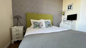 A neatly made bed with a grey knitted blanket, a green headboard, and two decorative pillows. A white bedside table with a lamp and a framed picture is to the left. A white dresser with a flat-screen TV and a vase with flowers is to the right.