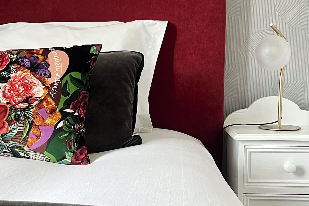 Un lit bien fait, avec une tête de lit rouge, du linge de lit blanc et un coussin à motifs floraux. On y trouve une table de chevet avec une lampe et un tiroir.